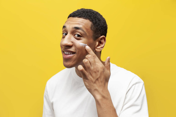 young african american guy in white t-shirt applies cream on his face on yellow isolated background, man uses facial cosmetics and takes care of his skin - Zdjęcie, obraz