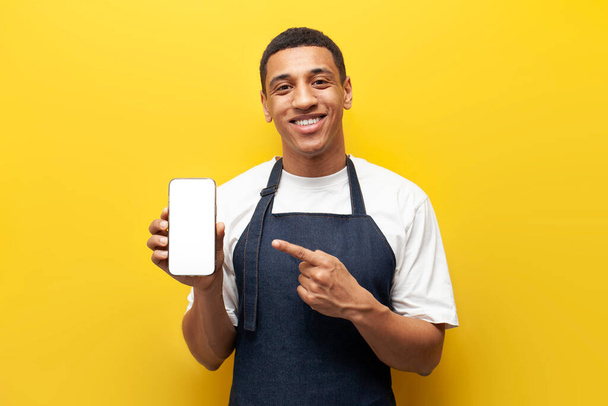 nuori Afrikkalainen amerikkalainen barista kaveri yhtenäinen pistettä tyhjä näyttö älypuhelimen keltaisella taustalla, jakelupalvelu työntekijä käyttää puhelinta, tarjoilija esiliina mainostaa verkossa järjestyksessä - Valokuva, kuva