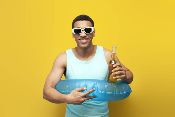 cheerful african american guy in sunglasses with inflatable swimming circle holds bottle of beer on yellow background, male tourist in summer at sea with alcohol, concept of travel and summer holidays - Foto, Imagen
