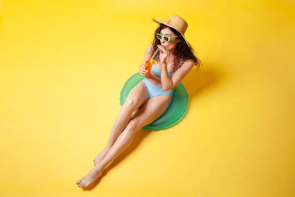 young girl in blue swimsuit sits on inflatable swimming ring holds orange alcoholic cocktail on yellow background, woman sunbathes on vacation in the summer - Foto, Bild