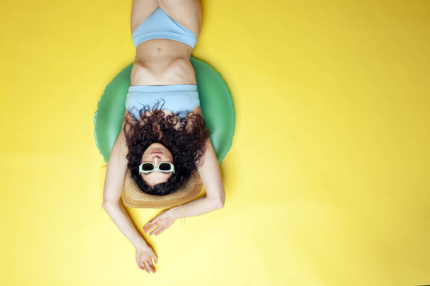 young girl in a blue swimsuit and sunglasses with an inflatable swimming circle lies on the beach and sunbathes in the summer, a woman on vacation at the sea on a yellow background, top view - Foto, Imagem