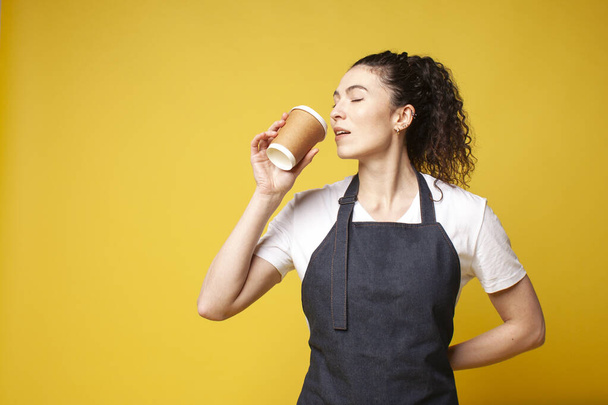 young barista girl in a denim apron drinks coffee on a yellow background, a portrait of a female waiter in uniform, copy space - Photo, Image