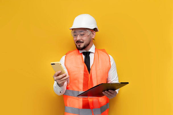 ázsiai civil mérnök egyenruhában használ okostelefon sárga elszigetelt háttér, férfi koreai kemény kalap és munkaruha gépel telefonon, építész munkavállaló - Fotó, kép