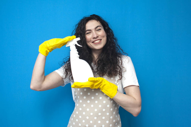 nő háziasszony kesztyű és kötény tartja mosószer a kezében a kék háttér, nő takarító hirdeti tisztító spray, takarítás szolgáltatás munkavállaló - Fotó, kép