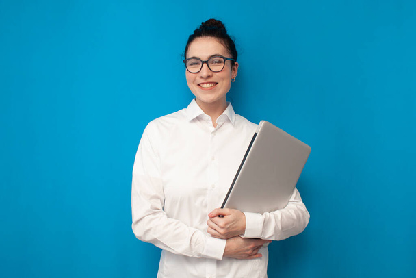 successful businesswoman in white shirt and glasses holds laptop on blue background and smiles, girl manager financier on colored background, portrait of office worker - Foto, afbeelding