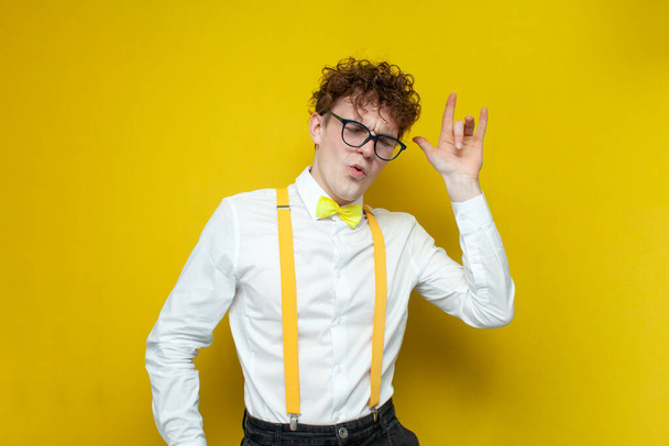 молодий впевнений веселий хлопець у святковому вбранні та окулярах радіє та танцює, носить студента у сорочці з бантом та підтягує руки на жовтому ізольованому фоні
 - Фото, зображення