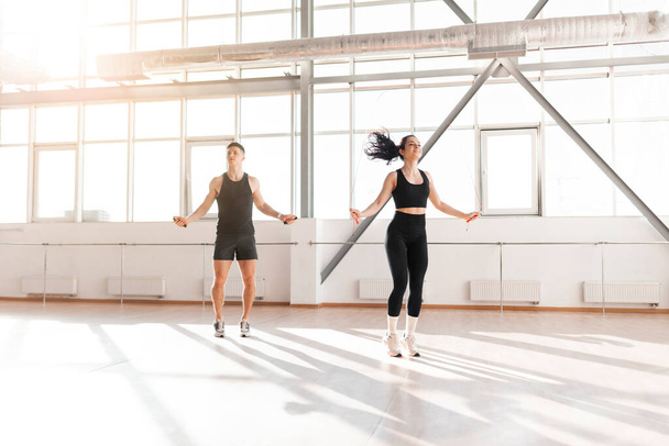 beautiful athletic couple in sportswear jumping rope in the gym in the morning, fitness girl and coach man go in for sports in training in bright room - Photo, Image