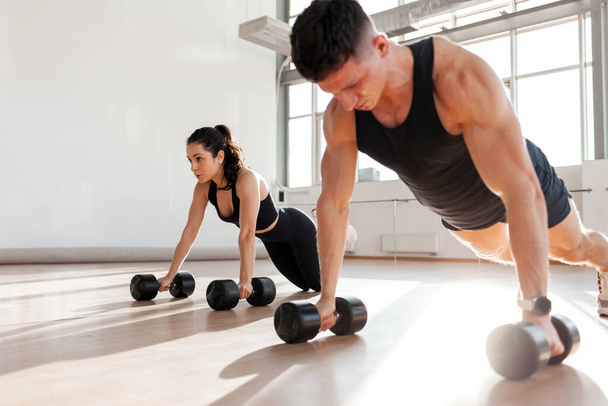 beautiful athletic couple in sportswear in training do push-ups with dumbbells in the fitness room in the morning, man trainer and fitness girl go in for sports in bright room - Foto, immagini