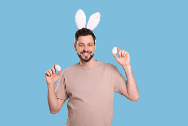 Happy man in bunny ears headband holding painted Easter eggs on turquoise background - Foto, Imagem