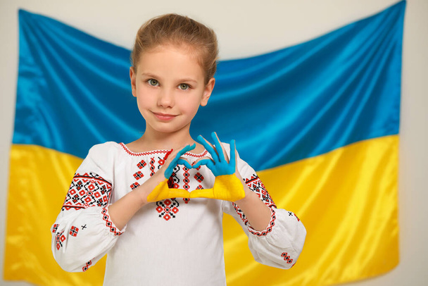 Little girl making heart with her painted hands near Ukrainian flag, space for text. Love Ukraine concept - Фото, изображение