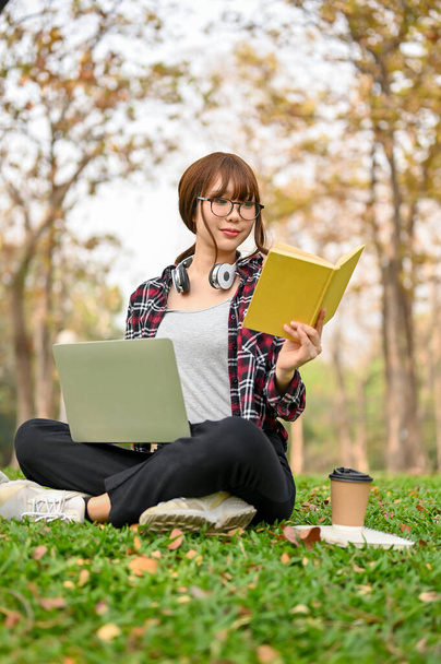 Πορτρέτο, Εστιασμένη και ελκυστική νεαρή Ασιάτισσα φοιτήτρια κάθεται στο γρασίδι στο πάρκο, διαβάζει ένα βιβλίο, και χρησιμοποιεί το laptop της για να διαχειριστεί τις σχολικές της εργασίες. έννοια uni-lifestyle - Φωτογραφία, εικόνα