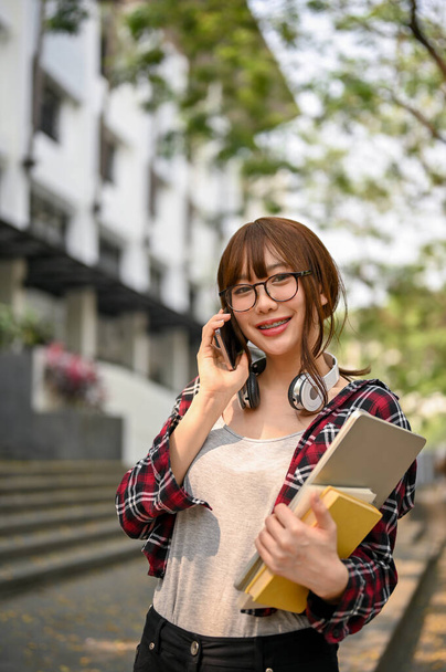 Portrait, Beautiful young Asian female college student in casual clothes and eyeglasses talking on the phone with her friend while standing outside of the campus building. - Fotó, kép