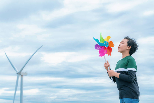 Progressive young asian boy playing with wind pinwheel toy in the wind turbine farm, green field over the hill. Green energy from renewable electric wind generator. Windmill in the countryside concept - Φωτογραφία, εικόνα
