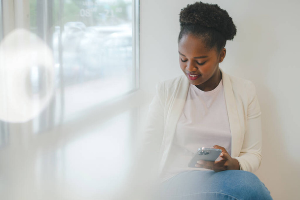 Vrolijk jong volwassen Afro-Amerikaans woma met afro krullend haar houden smartphone praten met een vriend aan de telefoon terwijl zittend in cafe vensterbank. Draadloos - Foto, afbeelding