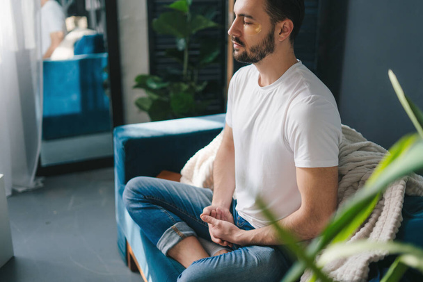 Photo of a young man doing yoga and meditating on the living room comfortable sofa. Morning relaxation and yoga practice concept - Foto, immagini