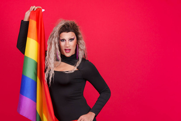 Трансгендер, одетый как трансгендер, смотрит в камеру с радужным флагом на фоне красного фона - Фото, изображение
