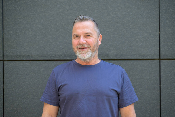 Een man met een baard van middelbare leeftijd die met een glimlach naar de camera kijkt terwijl hij tegen een muur poseert in een zomerT-shirt - Foto, afbeelding