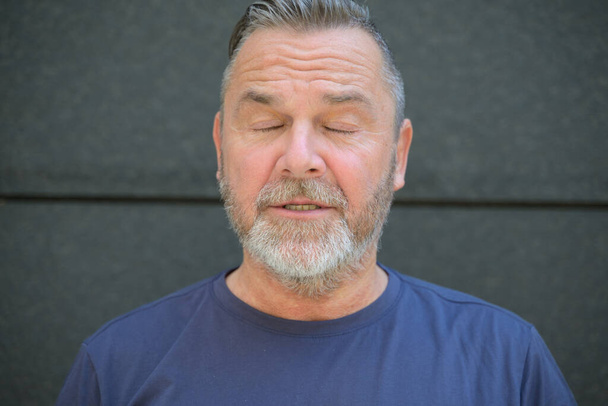 Середнього віку бородатий чоловік позує із закритими очима і роздвоєними губами на темній стіні
 - Фото, зображення