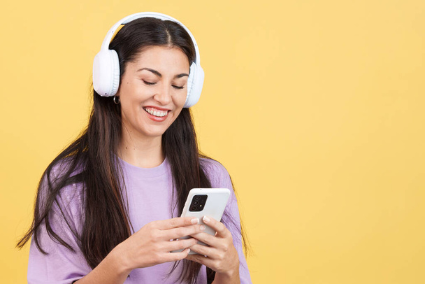 ヘッドフォンで音楽を聴く白人女性と黄色の背景を持つスタジオでのモバイル - 写真・画像