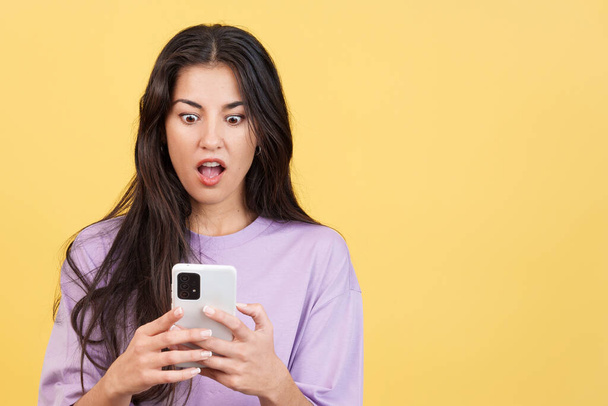 Zaskoczona kobieta patrząc na ekran telefonu komórkowego w studio z żółtym tle - Zdjęcie, obraz