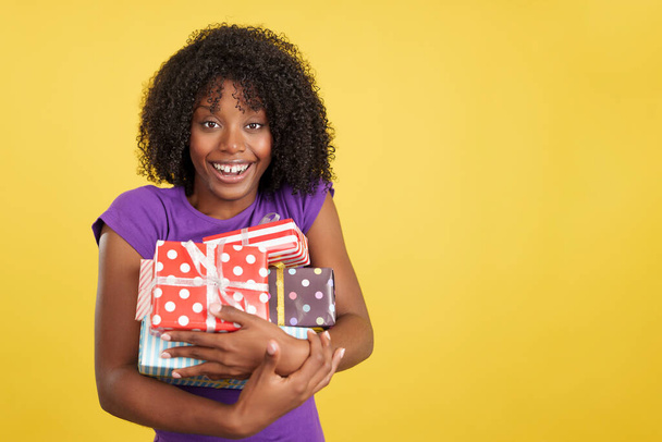 Glückliche Frau mit Afro-Frisur lächelt in die Kamera und hält Geschenke im Studio mit gelbem Hintergrund - Foto, Bild