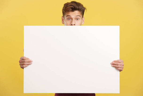 Überraschter Mann versteckt sich hinter einer leeren Platte im Atelier mit gelbem Hintergrund - Foto, Bild