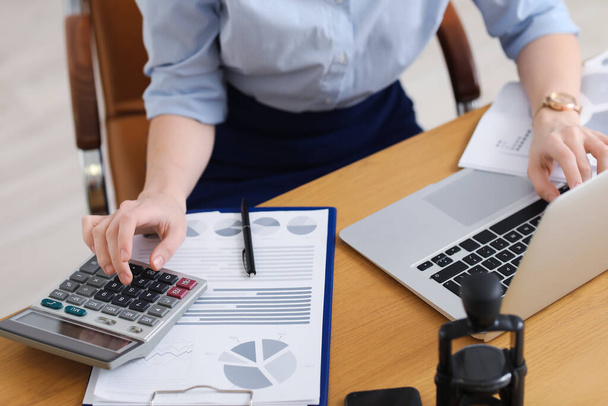 Женщина-бухгалтер работает с калькулятором и ноутбуком за столом в офисе, крупным планом - Фото, изображение