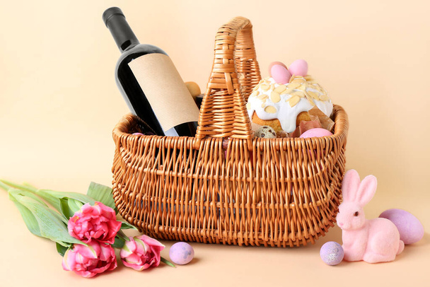 Корзина с пасхальными яйцами, торт, цветы тюльпана и бутылка вина на бежевом фоне - Фото, изображение