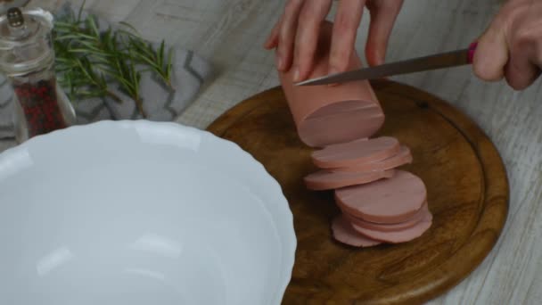 Cutting boiled sausage into circles with a kitchen knife on wooden cutting board. Close-up female hands slicing sausage on wooden cutting board with iron kitchen knife. Pepper grinder, rosemary sprig. - Materiał filmowy, wideo