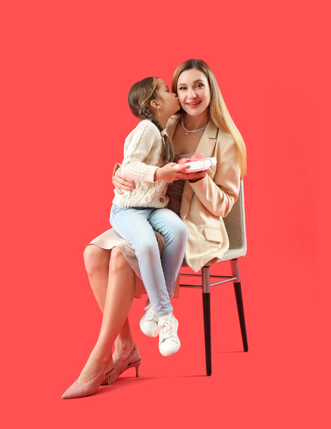 Χαριτωμένο κοριτσάκι με τη μητέρα της και το δώρο κάθεται σε κόκκινο φόντο - Φωτογραφία, εικόνα