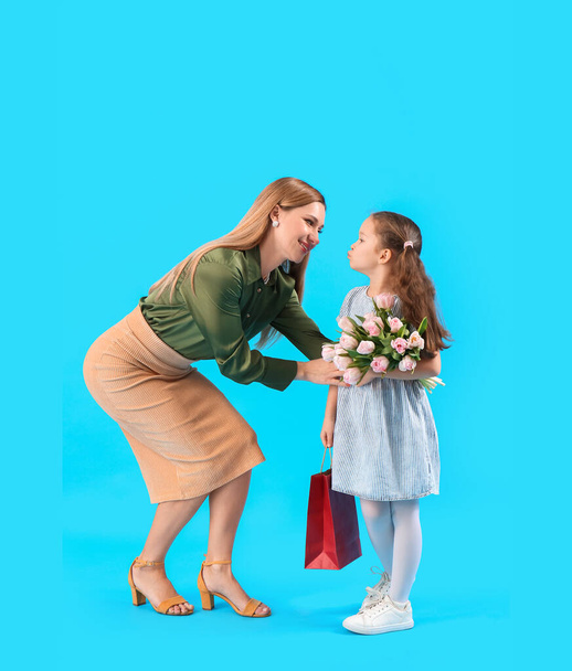 schattig klein meisje groeten haar moeder met tulpen en tas op blauwe achtergrond - Foto, afbeelding