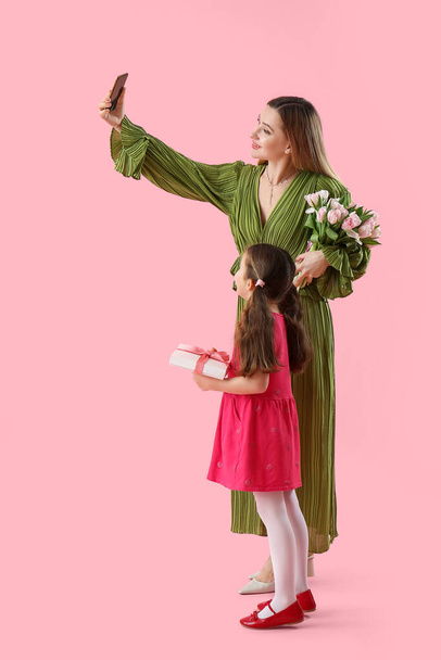 Linda niña con su madre y regalos tomando selfie sobre fondo rosa - Foto, imagen