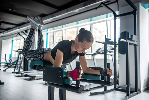 Chica deportiva muestra su cuerpo bien entrenado en el gimnasio donde hay muchas máquinas de ejercicio nuevas. Músculos bien desarrollados con entrenamiento de fuerza. - Foto, imagen