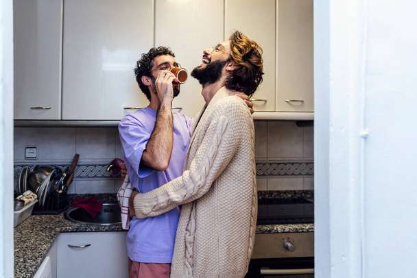 gay male couple hugging and laughing happy while having morning coffee in the kitchen, concept of home lifestyle and love between people of the same sex, copy space for text - Фото, изображение