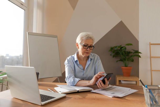 Αυτοπεποίθηση stylish Ευρωπαίος μεσήλικας ηλικιωμένη γυναίκα χρησιμοποιώντας smartphone στο χώρο εργασίας. Κομψό παλαιότερο ώριμο 60 ετών γκρίζα μαλλιά γυναίκα επιχειρηματίας με κινητό τηλέφωνο στο γραφείο. Αρχηγός αφεντικού που χρησιμοποιεί διαδικτυακές εφαρμογές - Φωτογραφία, εικόνα