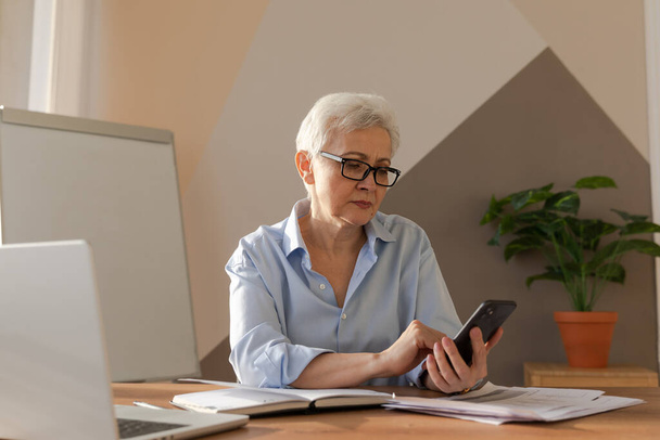 Confident stylish european middle aged senior woman using smartphone at workplace. Stylish older mature 60s gray haired lady businesswoman with cell phone in office. Boss leader using internet apps - Foto, Imagen