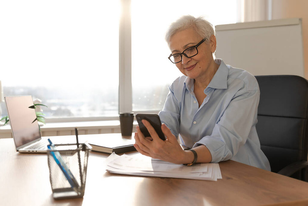 Confident stylish european middle aged senior woman using smartphone at workplace. Stylish older mature 60s gray haired lady businesswoman with cell phone in office. Boss leader using internet apps - 写真・画像