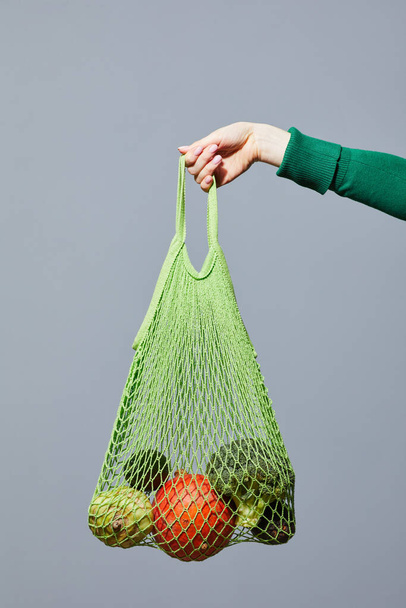 Minimal close up of hand holding eco shopping bag with vegetables against grey background, conscious consumption vegan concept - Valokuva, kuva