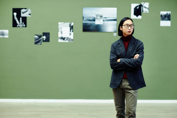 Horizontal medium long portrait of young Asian man wearing eyeglasses standing with arms crossed in modern art gallery looking at photos at exhibition - Φωτογραφία, εικόνα