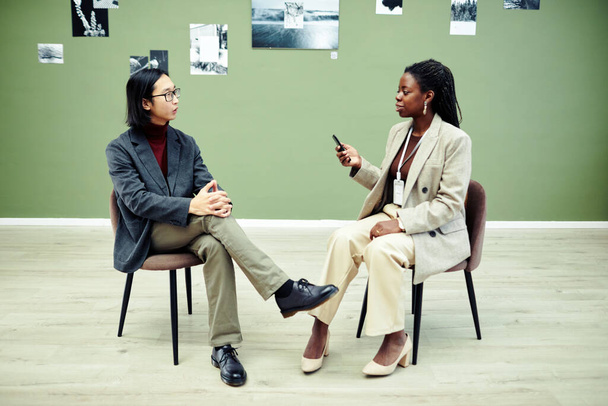 アフリカ系アメリカ人の女性が椅子に座ってボイスレコーダーを持っていて、現代の若いアジア人写真家にインタビューしています。 - 写真・画像