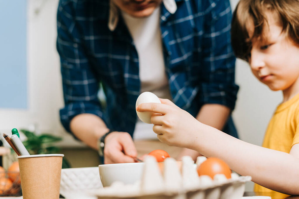 Ημέρα του Πάσχα. Αρσενικό Πατέρας και γιος ζωγραφική αυγά σε ξύλινο φόντο. Οικογένεια κάθεται σε μια κουζίνα. Προετοιμασία για το Πάσχα, δημιουργική σπιτική διακόσμηση. Παιδί παιδί αγόρι έχει τη διασκέδαση και τη ζωγραφική πασχαλινά αυγά - Φωτογραφία, εικόνα