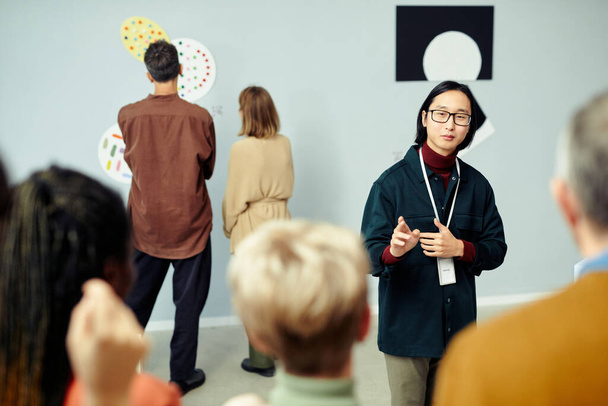 Sergide sergilenen çağdaş sanat eserleri hakkında konuşan bir grup turistin önünde duran genç Asyalı galerici - Fotoğraf, Görsel