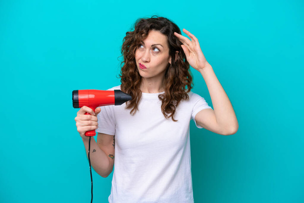 Young caucasian woman holding a hairdryer isolated on blue background having doubts and with confuse face expression - Foto, Bild