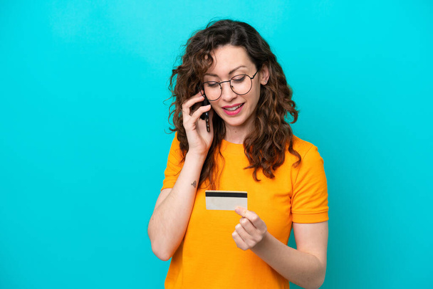 Νεαρή καυκάσια γυναίκα απομονωμένη σε μπλε φόντο αγοράζοντας με το κινητό με πιστωτική κάρτα - Φωτογραφία, εικόνα