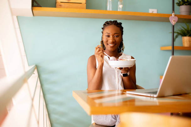 Mujer negra bastante joven desayunando saludable mientras trabaja en el ordenador portátil en la cafetería - Foto, Imagen