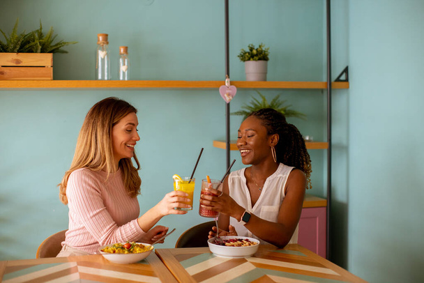 Dos mujeres negras y caucásicas bastante jóvenes pasándola bien, bebiendo jugos frescos y desayunando sano en la cafetería - Foto, imagen