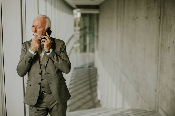 A senior business man stands in an office hallway, focused on his mobile phone. He is dressed in formal attire, exuding confidence and professionalism - Φωτογραφία, εικόνα
