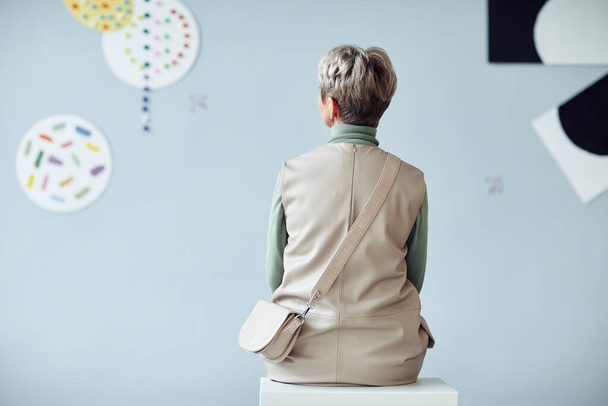 Задний вид неузнаваемой зрелой женщины, сидящей на стуле в галерее современного искусства и смотрящей на произведения искусства - Фото, изображение