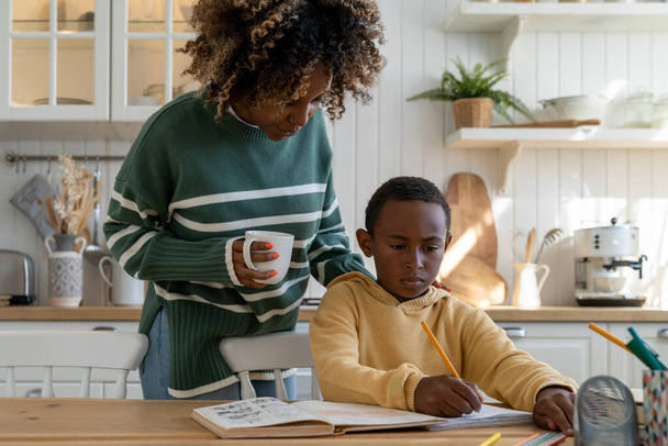 Afro-Amerikan anne çocukların ödevlerini kontrol ediyor, anne-baba öğrenme ile çocuğa destek oluyor, genç kadın anne okul çocuğunun başında durup oğlunun okulda başarılı olmasına yardım ediyor. Eğitimde ebeveyn rolü - Fotoğraf, Görsel
