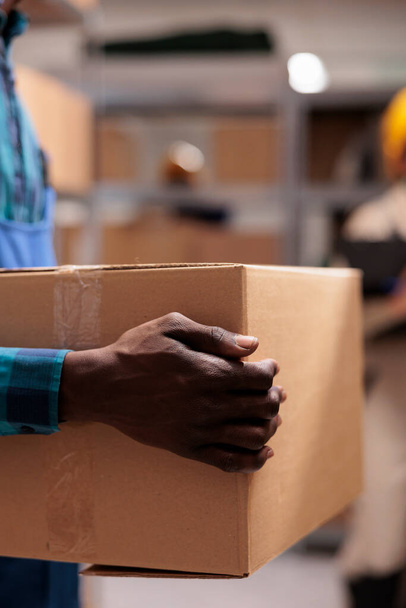 Υπάλληλος βιομηχανικής αποθήκης που κρατάει χάρτινο πακέτο έτοιμο για αποστολή. Αφρικανική αμερικανική ταχυδρομική υπηρεσία όπλων που μεταφέρουν δέμα πελάτη στο τμήμα διαλογής ταχυδρομείου κοντινό πλάνο αποθήκευσης - Φωτογραφία, εικόνα